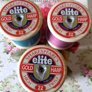 Op de afbeelding: Drie vintage Shakespeare's Elite katoenen garenspoelen. Elke spoel heeft een gouden, rode en blauwe label met de tekst "Shakespeare's Elite Gold High Cotton Thread Twist 22". De draadkleuren zijn blauw, roze en paars.