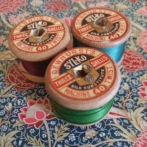 Puede incluir: Tres carretes de hilo de madera vintage con la inscripción "DEWHURST'S SYLKO SHELLS" y "SILK SUBSTITUTE MACHINE 40 TWIST". Los carretes están llenos de hilo burdeos, turquesa y verde, sobre un fondo floral.