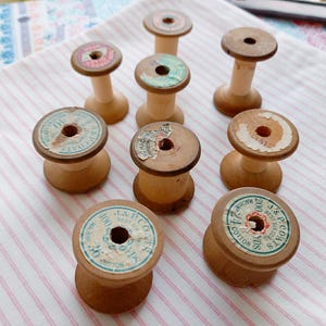 May include: A collection of vintage wooden spools with aged labels, some featuring text like "J & P Coats" and "Cotton." The spools are arranged on a pink and white striped fabric, showcasing their natural wood tones and historical charm.