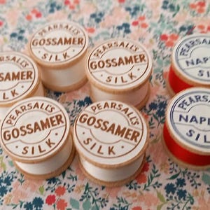 May include: A selection of vintage silk thread spools. The spools are white with wooden sides and feature the text "PEARSALL'S GOSSAMER SILK" and "PEARSALL'S NAPLES SILK" in a circular design. The thread colours are white and red.