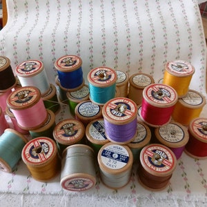May include: A collection of wooden spools of thread in various colours, including red, blue, green, yellow, and white. The spools are labelled with the brand name "Stikko" and other details about the thread.