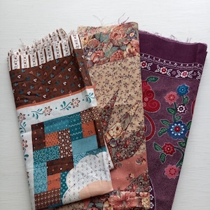 May include: Three folded fabric pieces with floral and patchwork patterns. The fabrics feature a mix of colours, including brown, teal, peach, and purple. The designs include floral prints, geometric patterns, and patchwork squares, suitable for quilting or crafting.