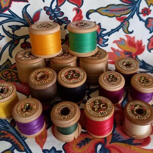 May include: A collection of vintage wooden spools of J&P Coats thread in various colours including yellow, orange, green, red, purple, and black. Each spool features a circular paper label on top with the brand name, thread weight, and yardage.