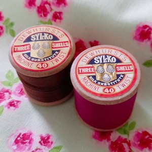Puede incluir: Dos carretes de hilo Sylko vintage. Uno es marrón oscuro, el otro magenta. Cada uno tiene un núcleo de madera y una etiqueta con "DEWHURST'S SYLKO THREE SHELLS MACHINE 40 TWIST". Ideal para costura y manualidades.