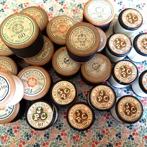 May include: A collection of vintage J&P Coats sewing thread spools. The spools are made of wood and cardboard, with labels featuring text and decorative elements. The thread colors include black, white, and various shades of brown. The spools are arranged on a floral patterned surface.