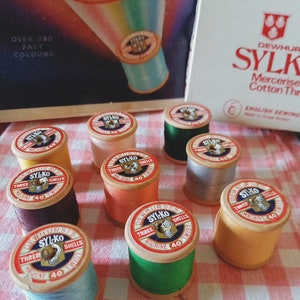 May include: A collection of ten spools of thread in various colours, including orange, green, blue, and brown. Each spool is labelled "Dewhurst's Sylko Three Shells Machine 40 Twist".
