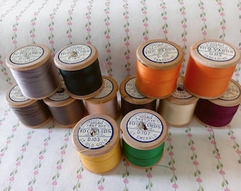 May include: A collection of 11 spools of thread, each with a wooden base and a paper label. The labels are all white with blue lettering and indicate the color of the thread. The colors include silver slate, dark green, tangerine, crimson, elephant, rose, old gold, light emerald, and cream.