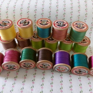 Op de afbeelding: Een verzameling van 18 vintage houten spoelen met garen in verschillende kleuren, waaronder rood, groen, paars, bruin en geel. Elke spoel heeft een label met de tekst "Sylko Triple Sheens 40".