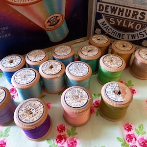 Könnte beinhalten: Eine Sammlung von Vintage Sylko Fast Dye Garnrollen in verschiedenen Farben wie Blau, Grün, Rosa und Lila. Jede Spule ist mit der Marke Sylko und dem Text "100 Yards" beschriftet. Die Spulen sind auf einem Stoff mit Blumenmuster angeordnet.