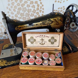 May include: Antique black Singer sewing machine with gold accents and a wooden box containing spools of thread. The box has the text "Clark & Co Paisley" and a nautical anchor logo.