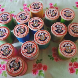May include: A collection of vintage sewing thread spools in various colours, including green, blue, red, and yellow. Each spool features a label with the text "Dewhursts Sylko Three Shells Machine 40 Twist".