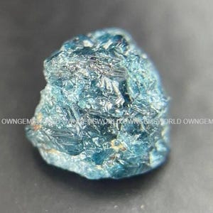 May include: A raw, uncut blue diamond crystal with a multifaceted, textured surface and deep azure tones. The stone displays a natural, irregular shape with sharp edges and reflective crystalline facets throughout its entire structure.