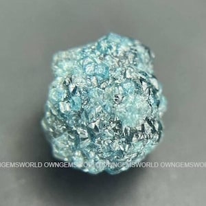 Peut inclure: Un cristal de diamant bleu brut et non taillé avec une surface texturée à facettes multiples. La pierre présente une teinte cyan vibrante avec des facettes métalliques réfléchissantes sur toute sa forme arrondie irrégulière. Le texte OWNGEMSWORLD est visible à la base.
