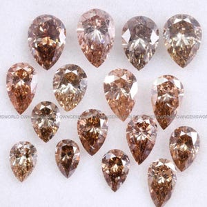 Puede incluir: Una colección de quince piedras preciosas sueltas en forma de pera en varios tonos de marrón, que van desde el champán claro hasta tonos chocolate intenso. Cada piedra presenta un corte facetado que refleja la luz, dispuesta sobre un fondo blanco.