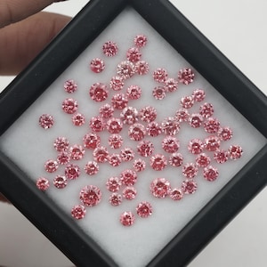 May include: A collection of numerous small, round, faceted pink gemstones arranged on a white foam insert within a square black display case. Each stone features a brilliant cut with multiple reflective surfaces that catch the light.