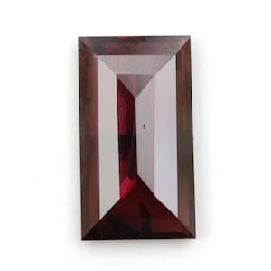 May include: A rectangular, faceted gemstone with a deep, translucent red color. The stone features a step-cut design with sharp, geometric edges and a polished surface that reflects light, creating distinct light and dark zones across its face.