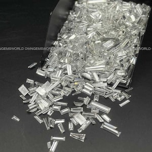 May include: A collection of clear, colorless, baguette-cut gemstones with rectangular and tapered shapes spilling out from a metal scoop. The stones have faceted surfaces that reflect light, and the text OWNGEMSWORLD is visible in the background.