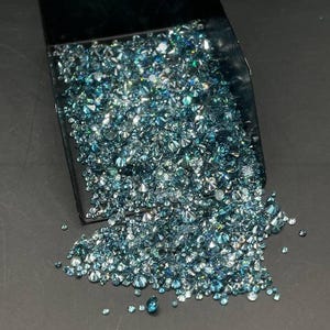 以下が含まれることがあります： 黒いプラスチック製のスコップに入った、小さなファセットカットの丸い青い宝石の山。石は様々な色合いのティールやシアン色をしており、一部がスコップから下の面にこぼれ落ち、光を反射して輝いています。