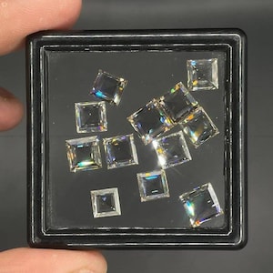 May include: A collection of eleven clear, square-cut gemstones arranged inside a square black display case. Each stone features a faceted surface that reflects light, creating a prismatic effect with subtle rainbow-coloured flashes across the facets.