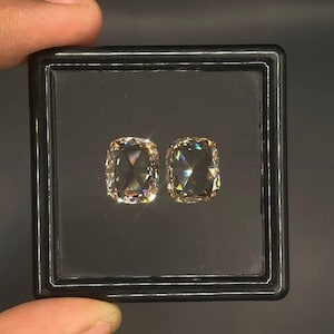 Puede incluir: Dos piedras preciosas de color champán, con talla cojín y superficies facetadas, se muestran una al lado de la otra dentro de un estuche cuadrado de plástico negro con tapa transparente, reflejando la luz con un brillo intenso.