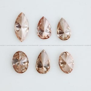 May include: A collection of six loose faceted gemstones in a light champagne or peach-toned color. The set includes one marquise cut, three pear-shaped stones, and two oval-cut stones, all featuring intricate geometric facets that reflect light.