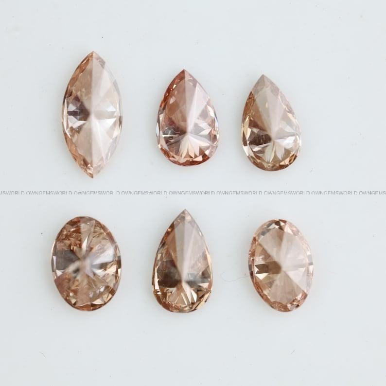 May include: A collection of six loose faceted gemstones in a light champagne or peach-toned color. The set includes one marquise cut, three pear-shaped stones, and two oval-cut stones, all featuring intricate geometric facets that reflect light.