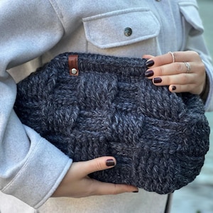 May include: A dark gray knitted clutch bag with a brown leather strap and buckle closure. The bag is held in a person's hand.