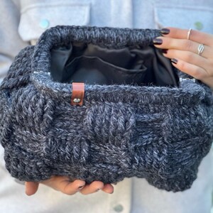 May include: A gray knitted bag with a brown leather handle. The bag is made of a thick, textured yarn and has a woven design.