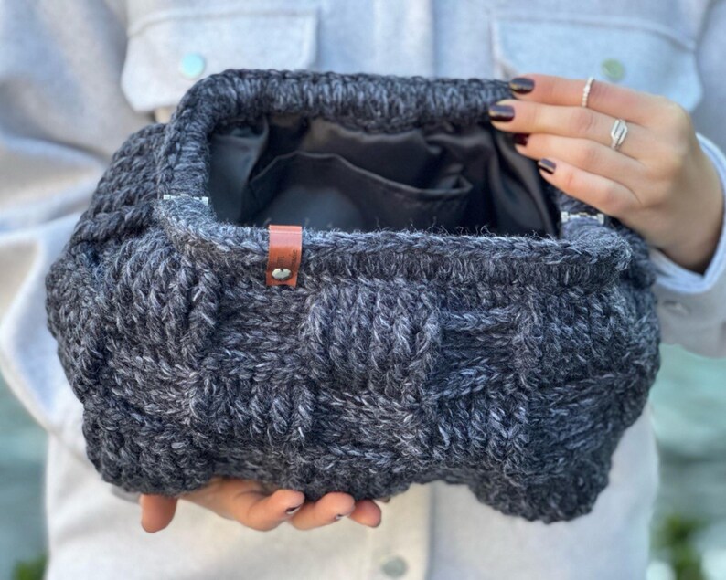 May include: A gray knitted bag with a brown leather handle. The bag is made of a thick, textured yarn and has a woven design.