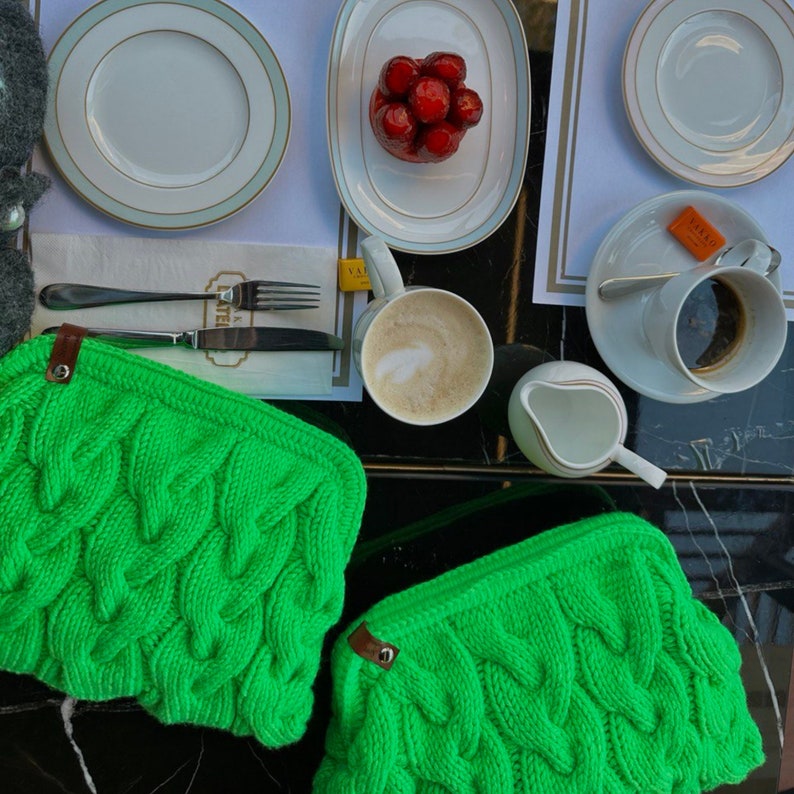 May include: Two bright green knitted bags with a cable knit pattern. The bags are on a dark surface with a cup of coffee and a plate of dessert in the background.