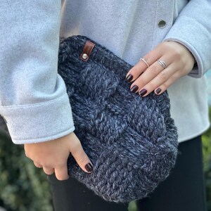 May include: A dark gray knitted clutch bag with a brown leather tag. The bag has a woven pattern and is held by a person wearing a gray sweater.