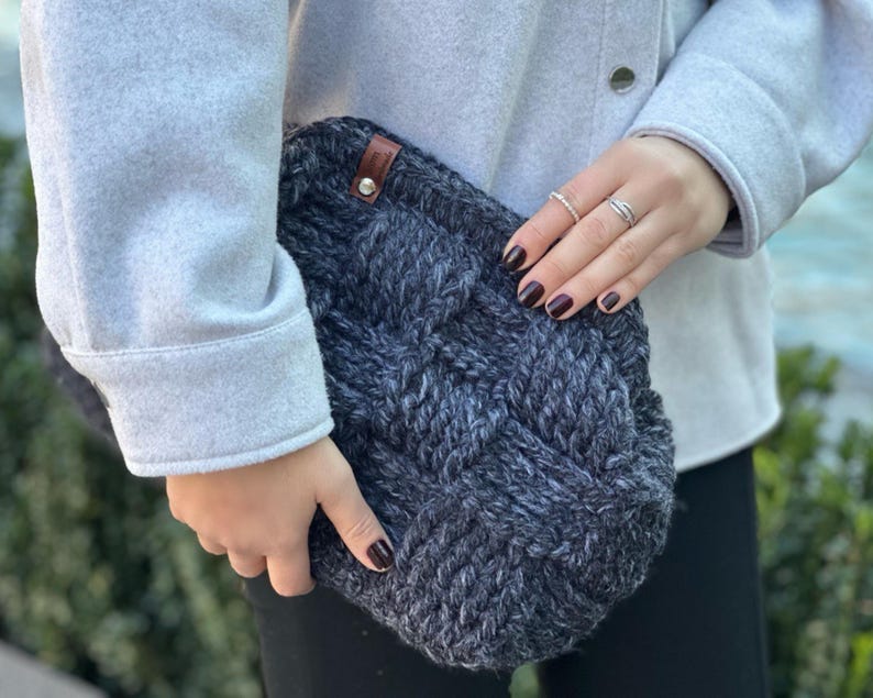May include: A dark gray knitted clutch bag with a brown leather tag. The bag has a woven pattern and is held by a person wearing a gray sweater.