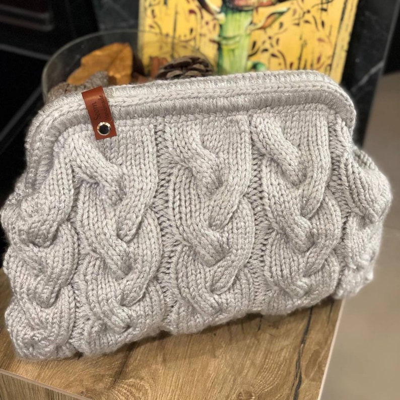 May include: A gray knitted clutch bag with a cable knit pattern. The bag has a brown leather tag with the word "Sofia" on it.