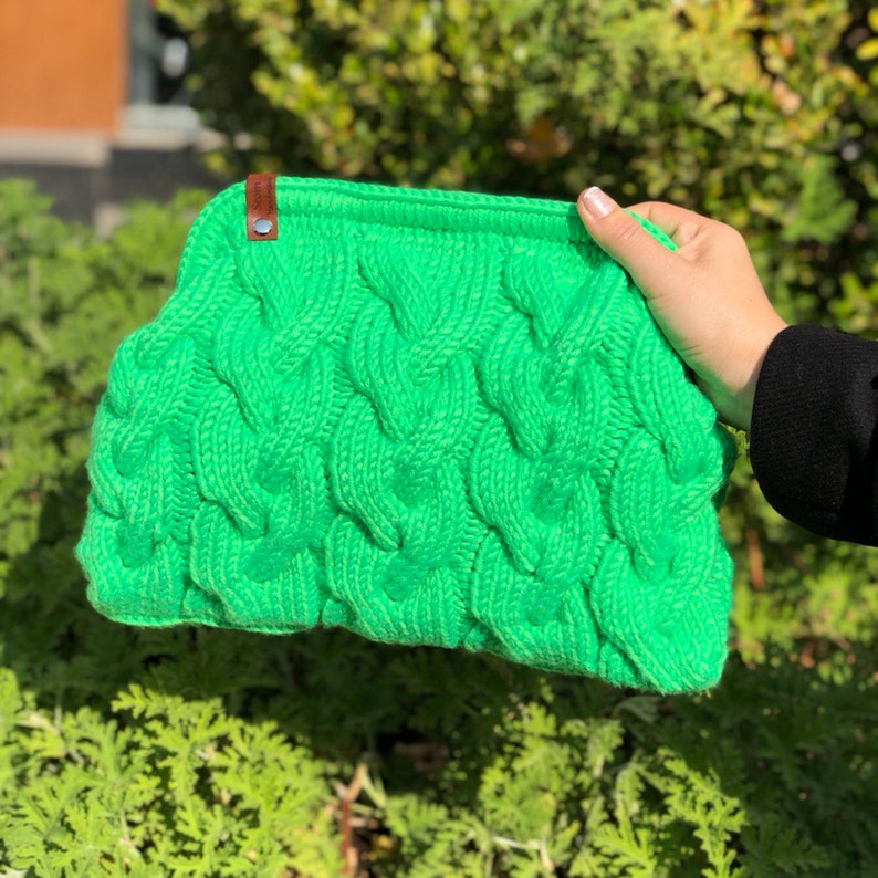 May include: A bright green knitted clutch bag with a cable knit pattern. The bag has a brown leather tag.