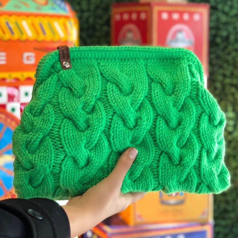 May include: A bright green knitted clutch bag with a cable knit pattern. The bag has a brown leather tag with the word "Sorm" on it.