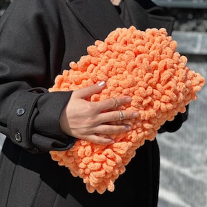 May include: A soft, peach-colored pillow made of fluffy yarn. The pillow is held in a person's hand.