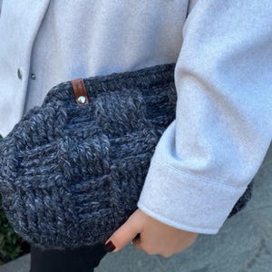May include: A gray knitted tote bag with a brown leather handle. The bag has a woven texture and is held by a person wearing a light gray coat.