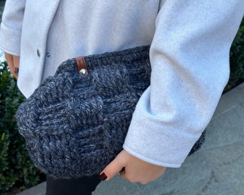 May include: A gray knitted tote bag with a brown leather handle. The bag has a woven texture and is held by a person wearing a light gray coat.