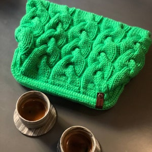 May include: A bright green knitted cable knit throw blanket with a brown leather tag that says "Stone Island". Two black and white ceramic mugs with coffee are on a gray surface.