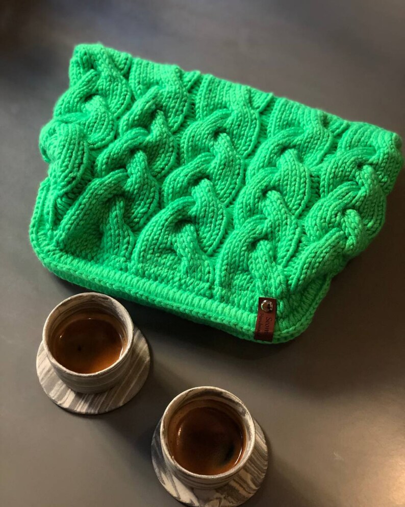 May include: A bright green knitted cable knit throw blanket with a brown leather tag that says "Stone Island". Two black and white ceramic mugs with coffee are on a gray surface.