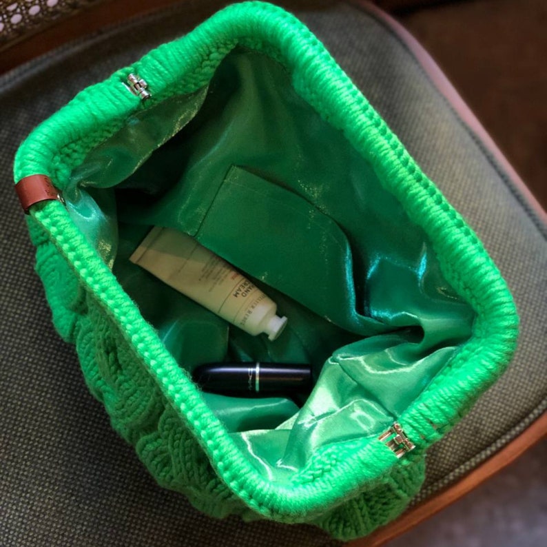 May include: A bright green knitted bag with a satin lining. The bag is open and has a tube of hand cream and a black lipstick inside.