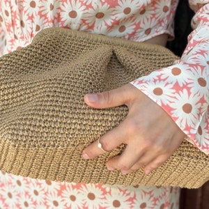 May include: A tan woven clutch bag with a textured surface. The bag is held in a hand wearing a gold ring.