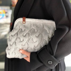 May include: A gray knitted clutch bag with a cable knit pattern and a brown leather tag. The bag is held by a person wearing a black coat.