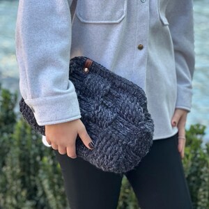 May include: A gray and black knitted clutch bag with a brown leather accent. The bag has a braided design and is held by a person wearing a gray shirt and black pants.