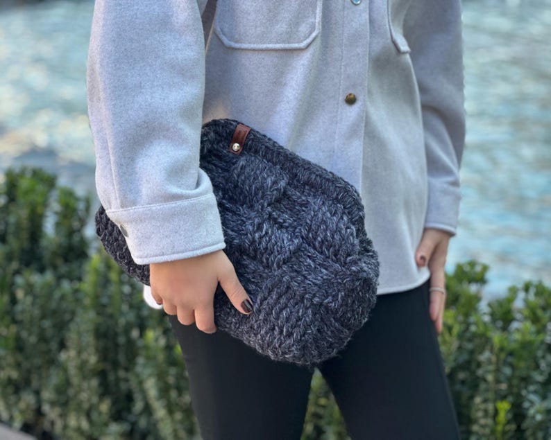 May include: A gray and black knitted clutch bag with a brown leather accent. The bag has a braided design and is held by a person wearing a gray shirt and black pants.