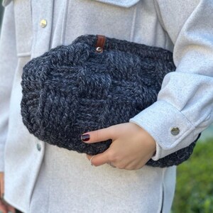May include: A gray knitted tote bag with a brown leather handle. The bag has a woven texture and is held by a person.
