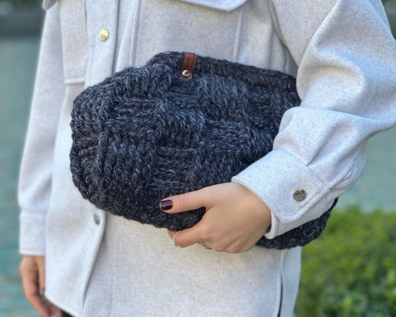 May include: A gray knitted tote bag with a brown leather handle. The bag has a woven texture and is held by a person.
