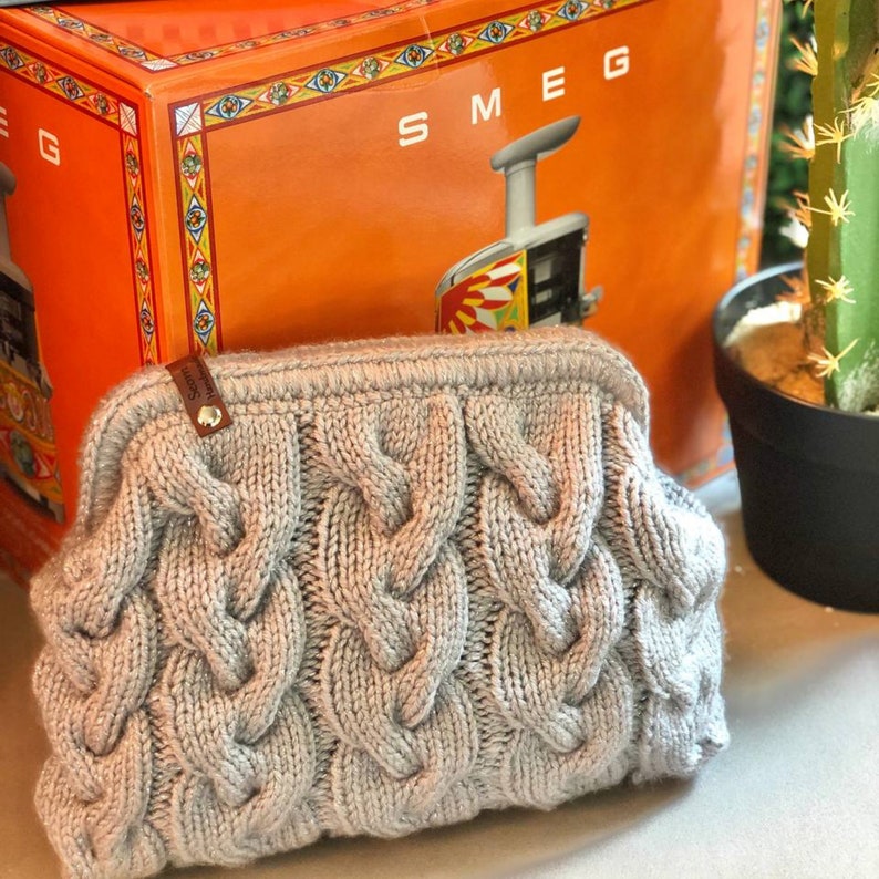 May include: A gray knitted clutch bag with a cable knit pattern. The bag has a small leather tag with the word "Sgom" on it.