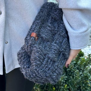 May include: A gray knitted tote bag with a brown leather strap. The bag is made of a thick, textured yarn and has a slouchy, casual look.