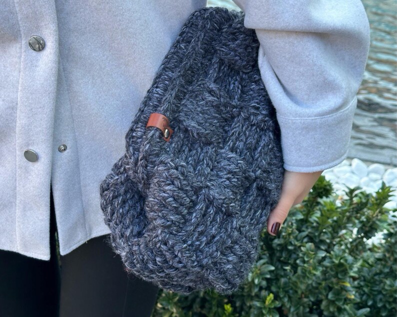 May include: A gray knitted tote bag with a brown leather strap. The bag is made of a thick, textured yarn and has a slouchy, casual look.